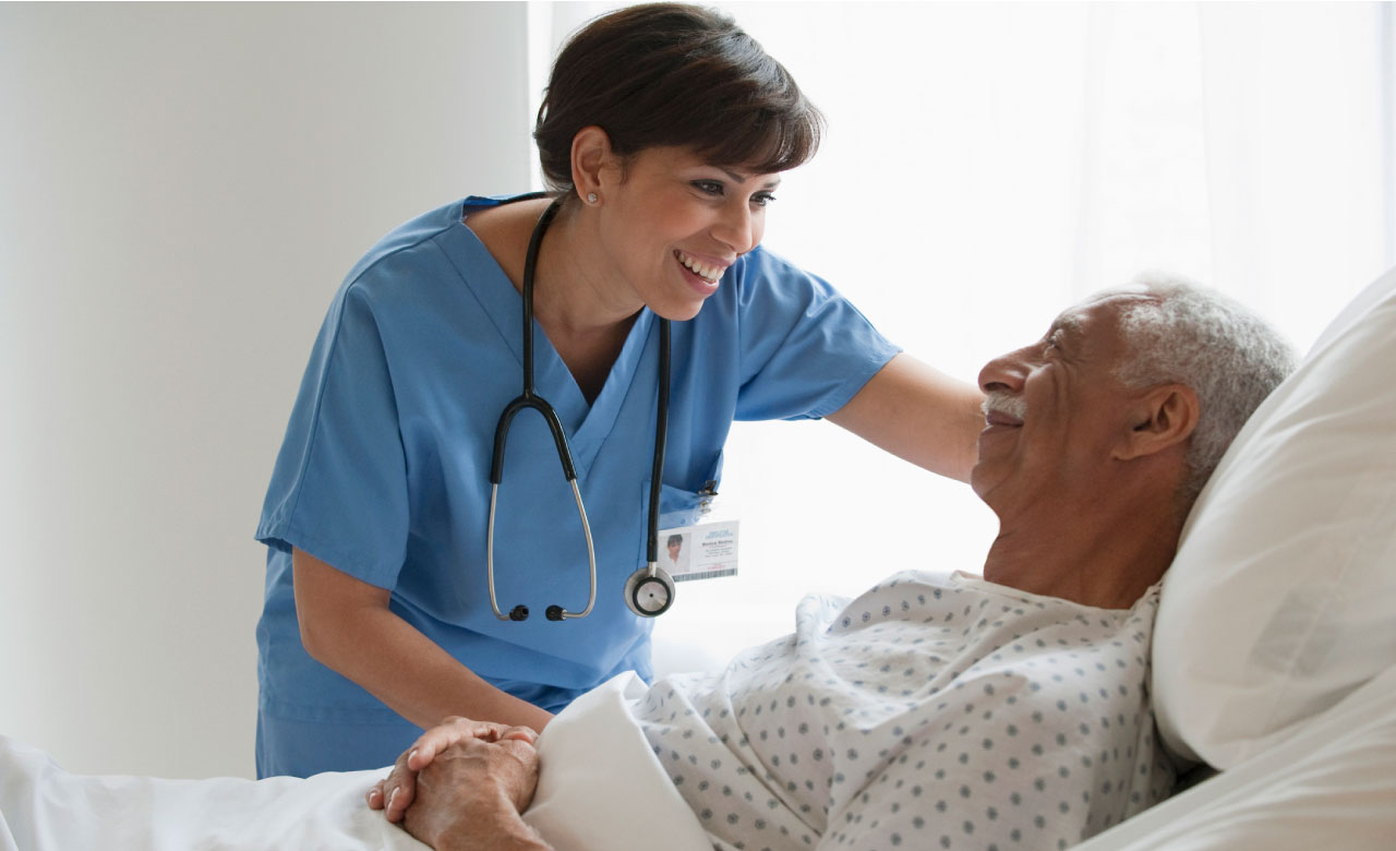 A medical professional wearing scrubs and a stethoscope stands over the bed of an elderly patient in a supportive and welcoming manner. The elderly patient is smiling.  