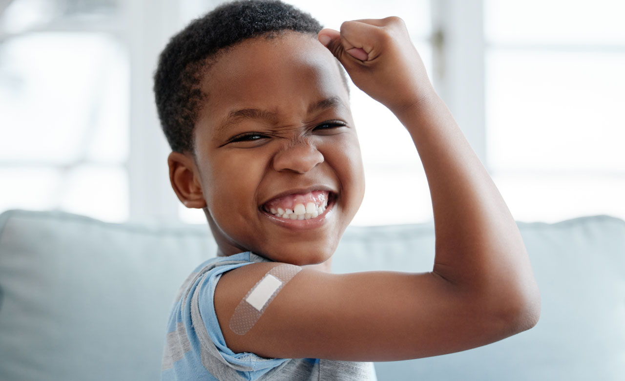 Young black boy grins and flexes his arm at the camera, showing off an immunization bandage. 